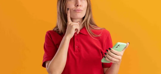 thoughtful young pretty woman puts finger chin holds phone isolated orange wall_compressed 540x250